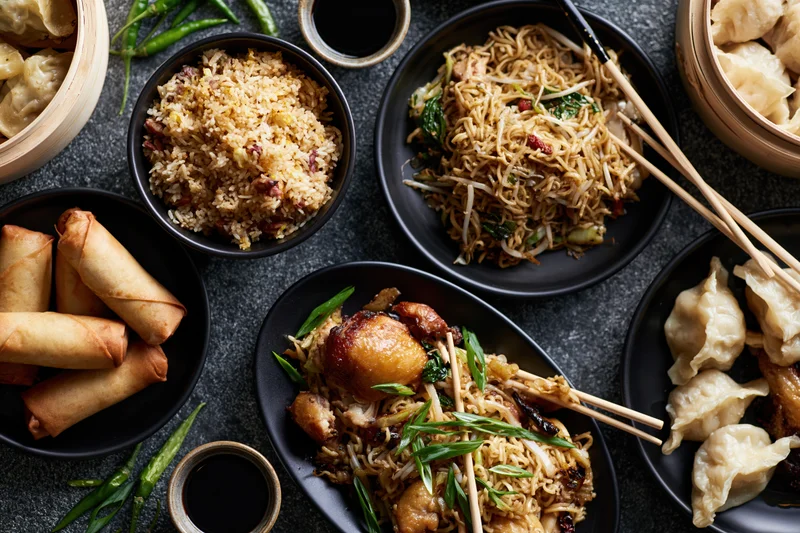 Overhead flat-lay of assorted chinese dishes including stir-fried noodles, fried rice, spring rolls, dumplings, and soy sauce on a dark surface
