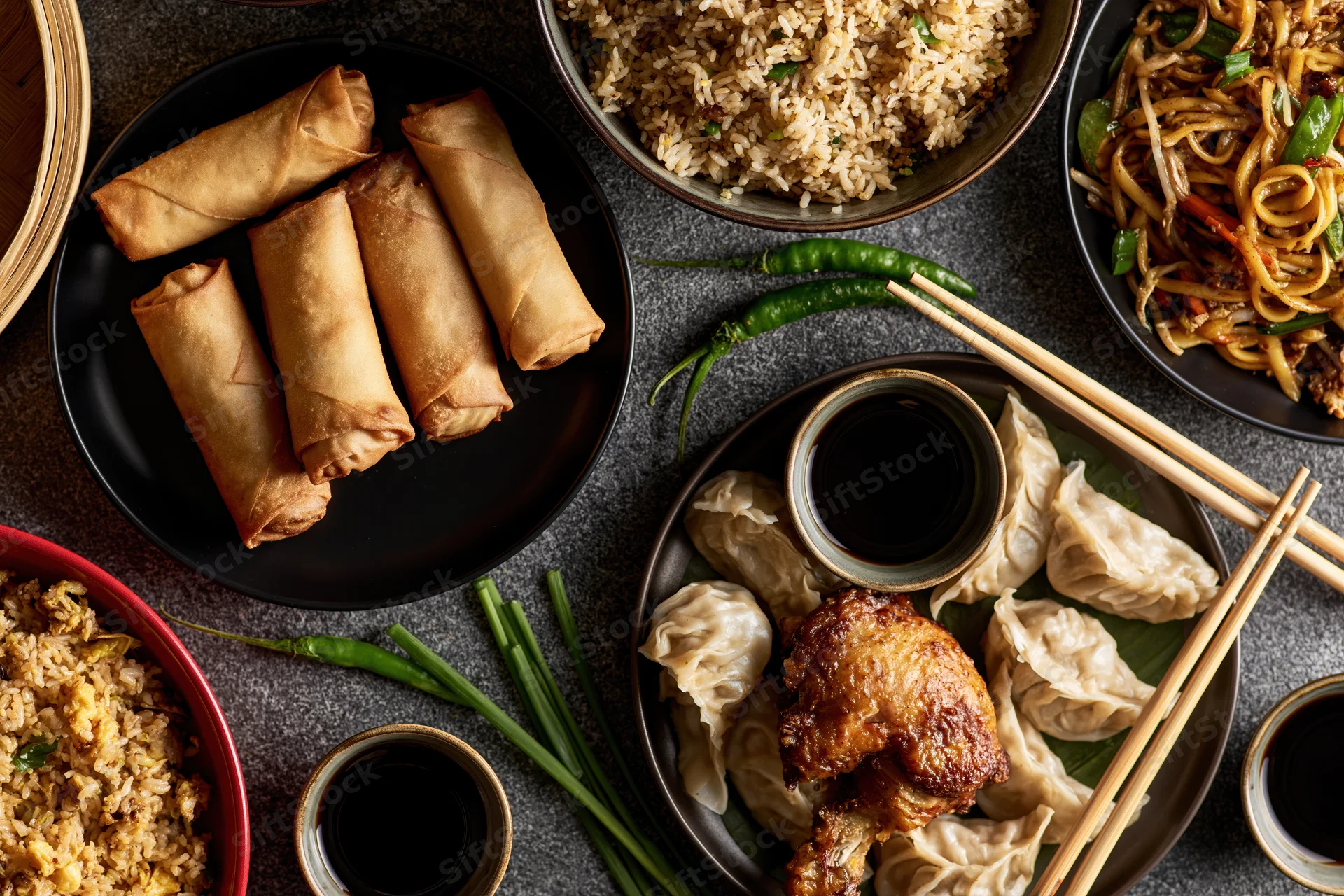 Overhead flat-lay of assorted chinese dishes including spring rolls, dumplings, fried rice, noodles, and soy sauce arranged on a dark stone surface with chopsticks and green chili peppers