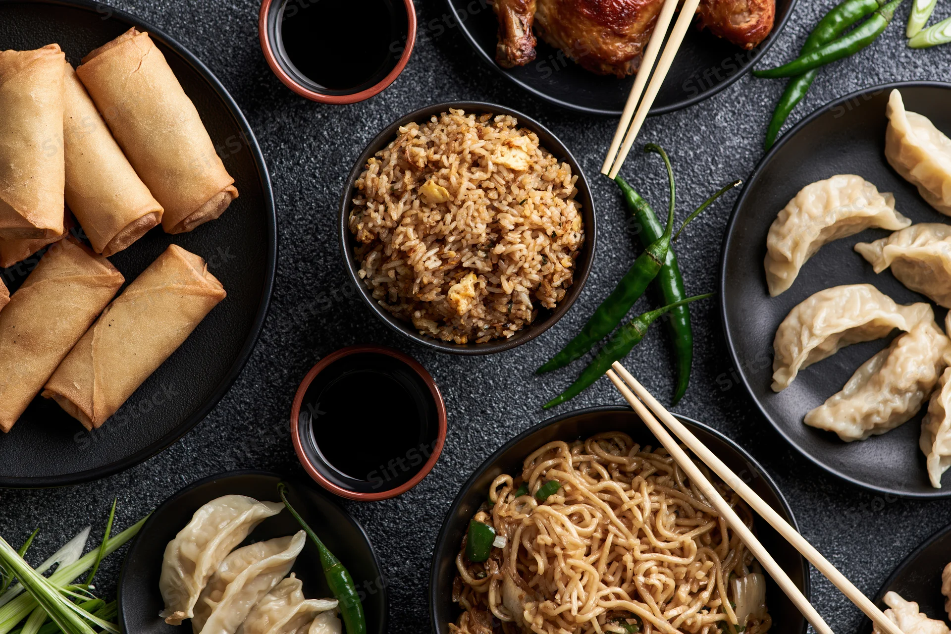 Overhead flat-lay of assorted chinese dishes including spring rolls, fried rice, noodles, dumplings, soy sauce, and chili peppers arranged on black plates and bowls on a dark surface