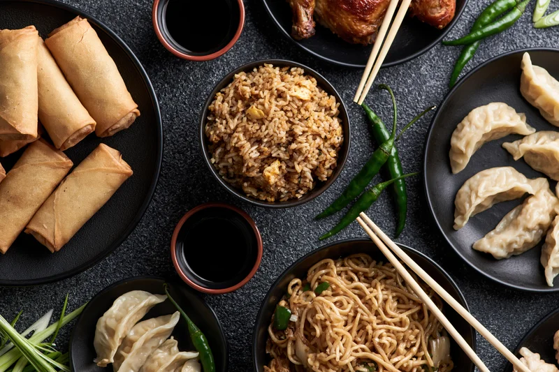 Overhead flat-lay of assorted chinese dishes including spring rolls, fried rice, noodles, dumplings, soy sauce, and chili peppers arranged on black plates and bowls on a dark surface