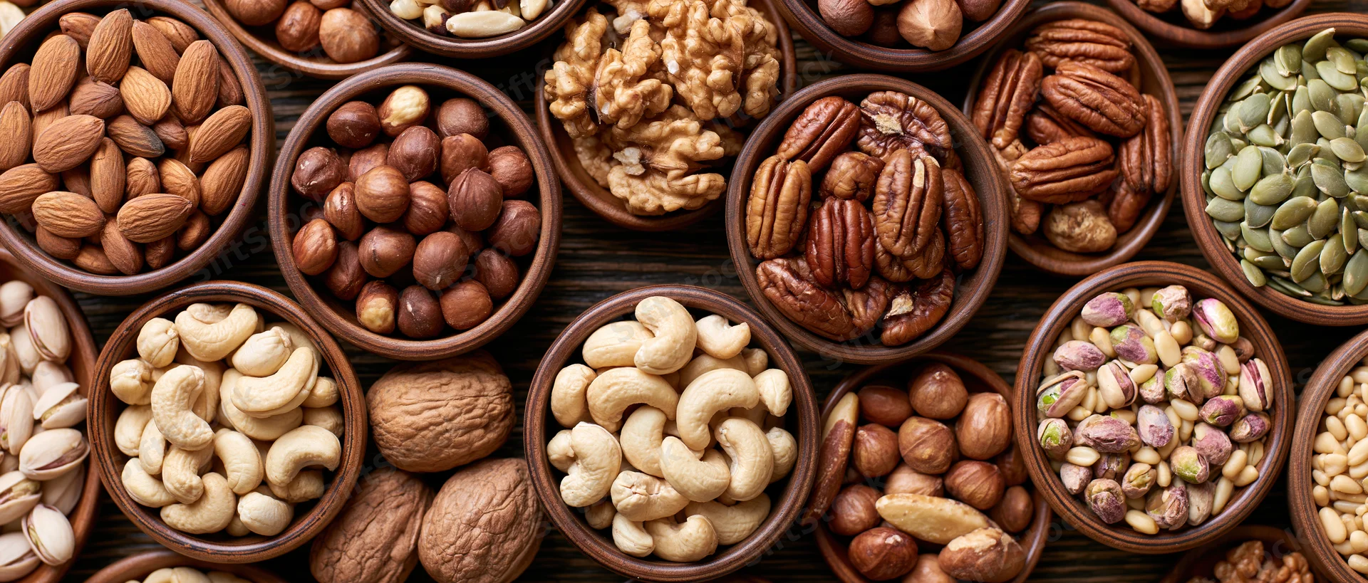 Assorted nuts and seeds in wooden bowls arranged in overhead flat lay on dark surface