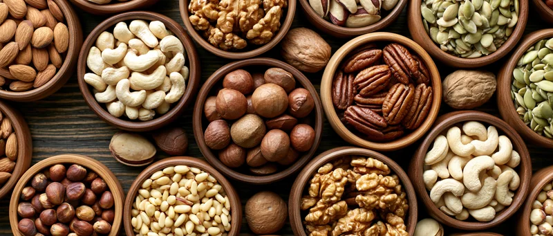 Assorted nuts and seeds in wooden bowls arranged in overhead flat lay on dark wood surface
