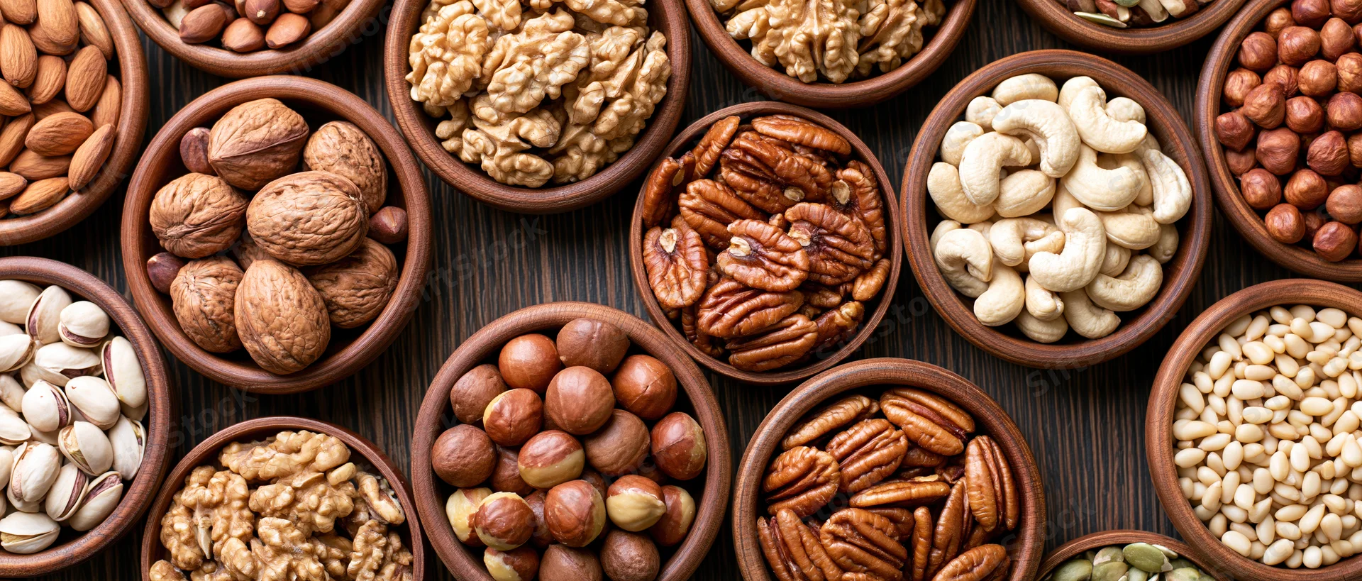 Assorted nuts and seeds in wooden bowls arranged in flat lay on dark wood surface