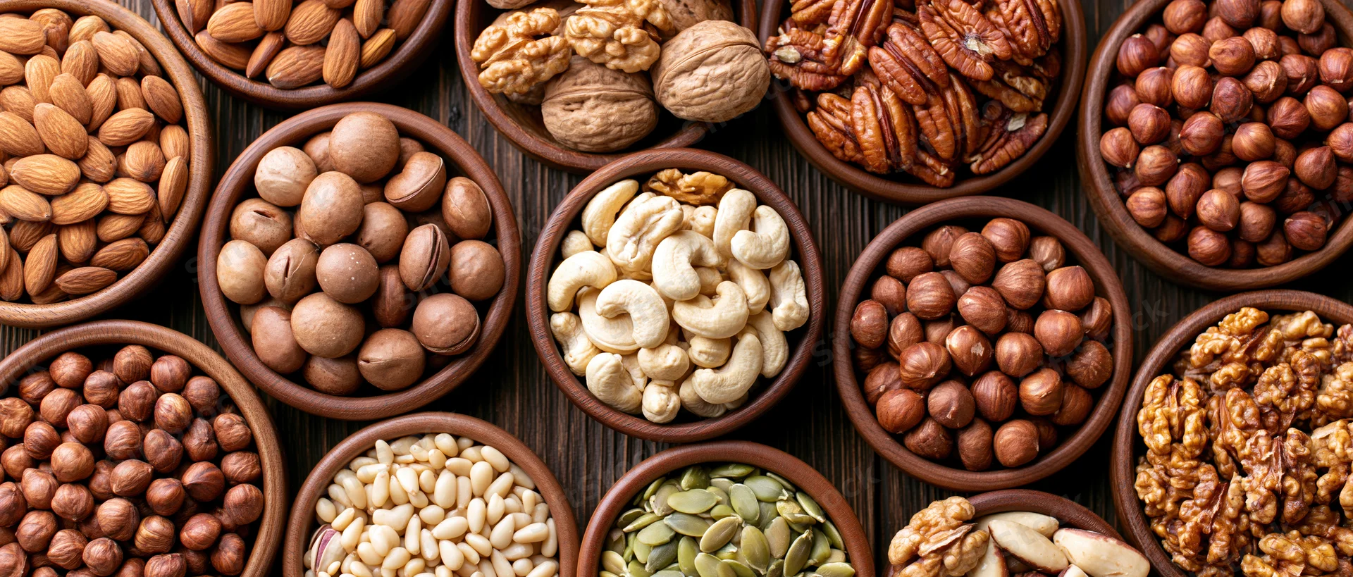 Assorted nuts and seeds in wooden bowls arranged in overhead flat lay on dark wood surface