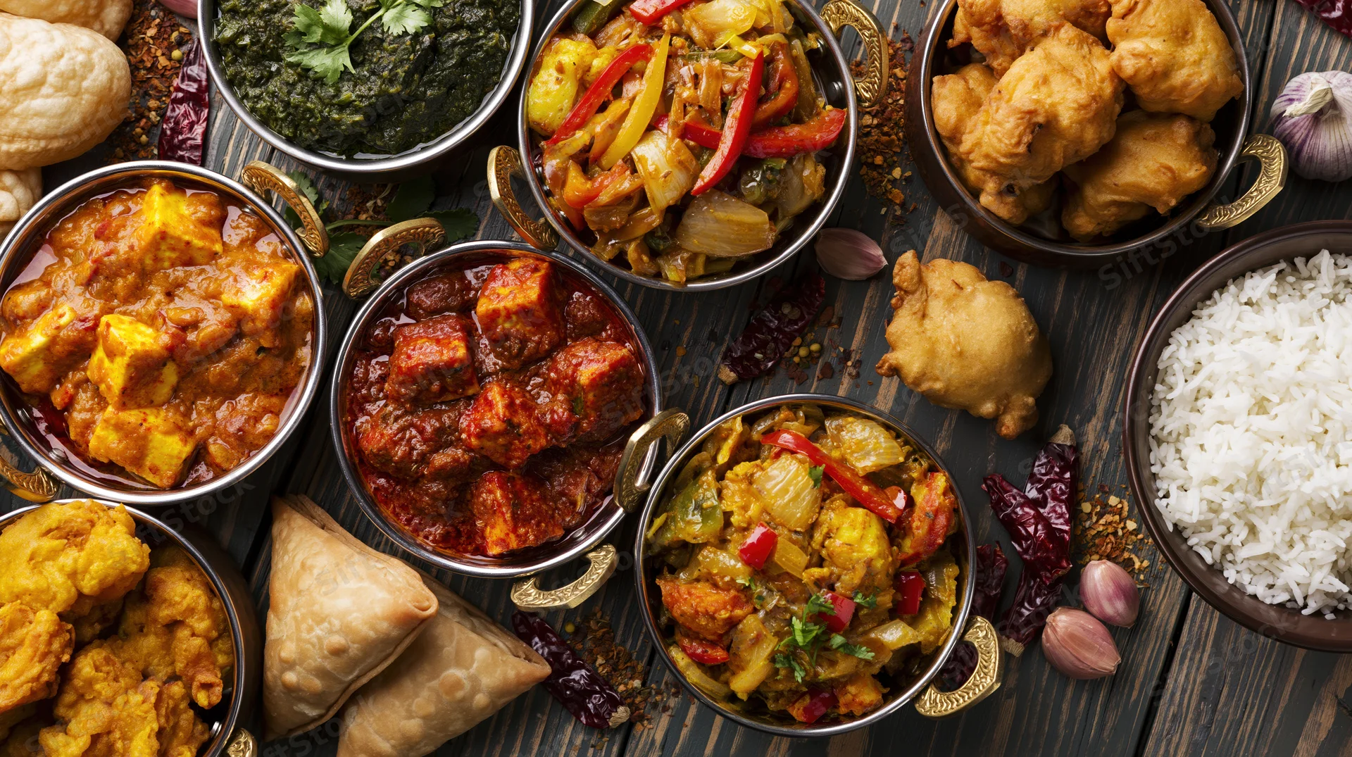Overhead flat-lay of multiple Indian dishes arranged on a wooden table with bowls of curry, rice, samosas, naan, and spices