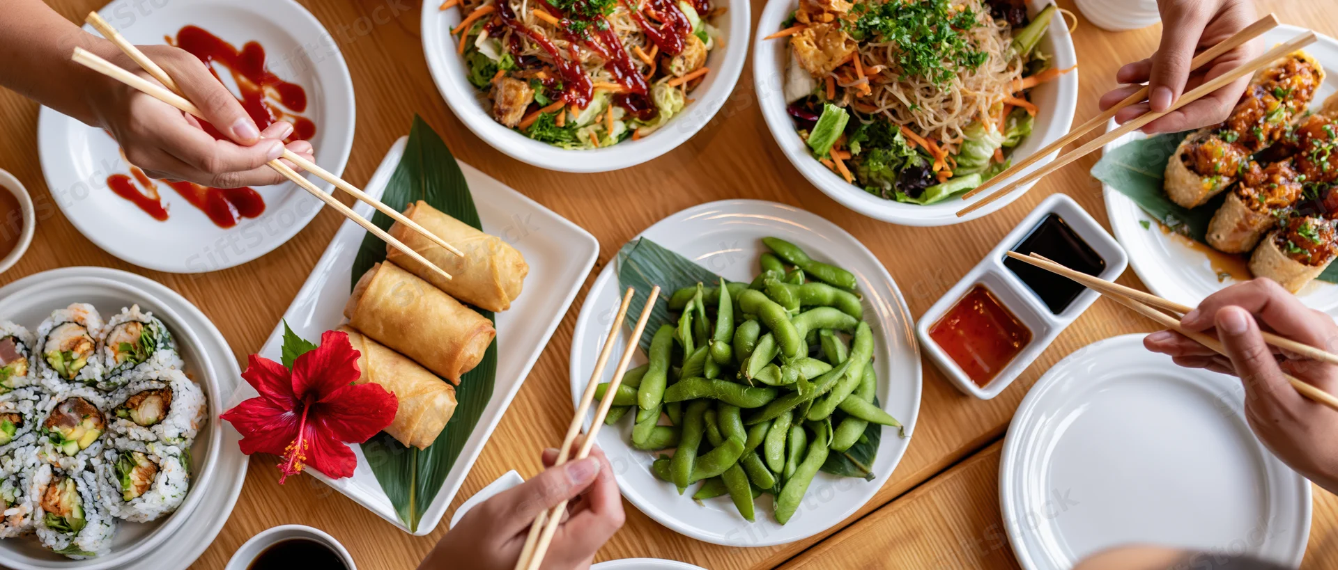 People sharing a spread of sushi, spring rolls, edamame and sauces on a wooden table with chopsticks
