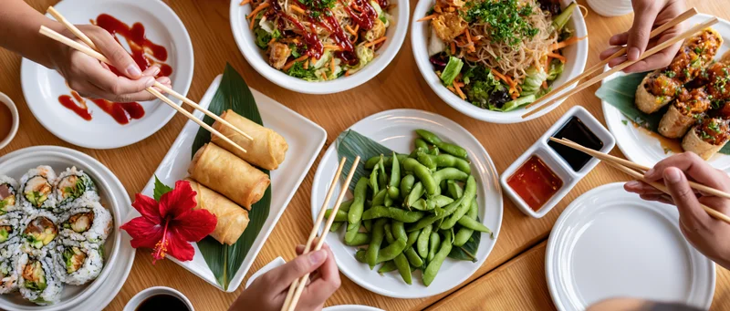 People sharing a spread of sushi, spring rolls, edamame and sauces on a wooden table with chopsticks