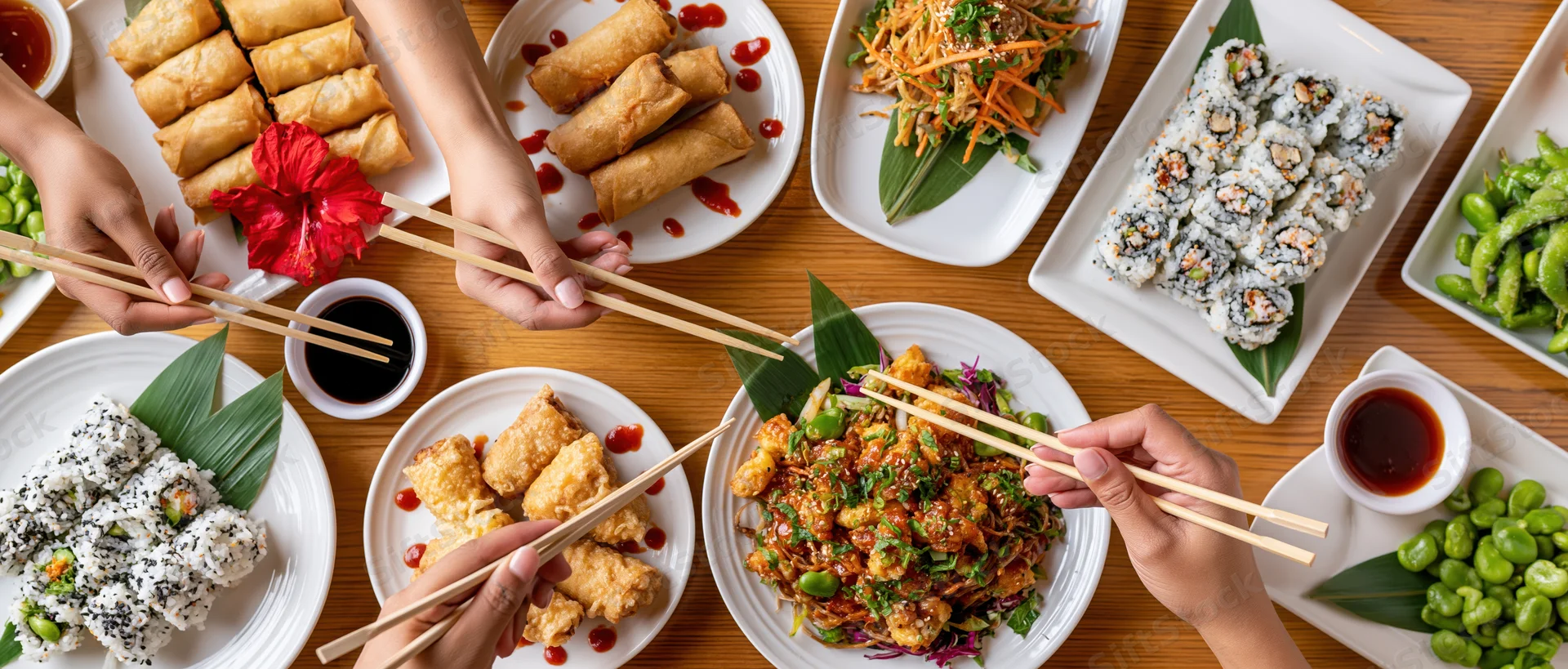 People sharing a spread of sushi, spring rolls, edamame and stir-fry on a wooden table with chopsticks and soy sauce