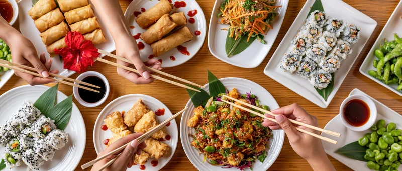 People sharing a spread of sushi, spring rolls, edamame and stir-fry on a wooden table with chopsticks and soy sauce