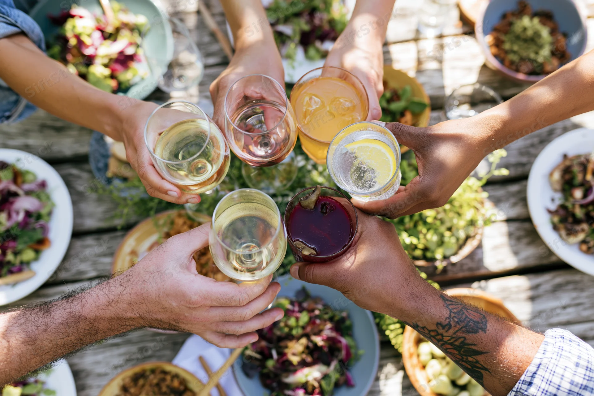 Diverse hands clinking glasses with drinks and food on a wooden table outdoors