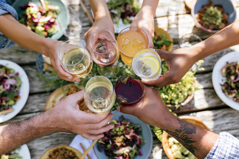 Diverse hands clinking glasses with drinks and food on a wooden table outdoors