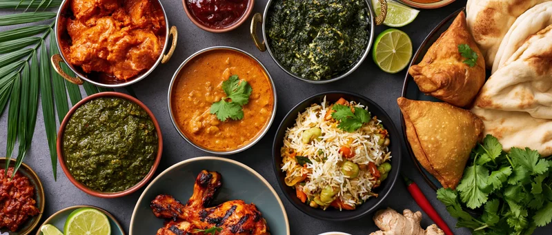 Overhead flat-lay of indian dishes including curry, rice, naan, samosas and tandoori chicken on dark surface
