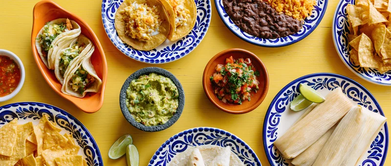Overhead flat-lay of mexican dishes including tacos, guacamole, salsa, beans, rice, chips, and tamales on a yellow table