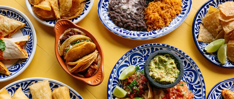 Overhead flat-lay of mexican dishes including tacos, guacamole, rice, beans, and chips arranged on a yellow table with blue and white patterned plates