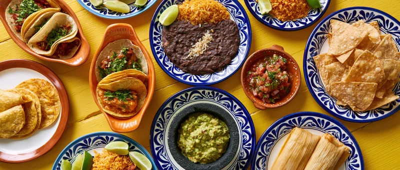 Overhead flat-lay of mexican dishes including tacos, tamales, guacamole, and chips arranged on a yellow table