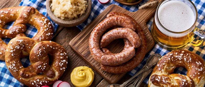 Overhead flat lay of bavarian oktoberfest food spread with beer, pretzels, sausage, and sauerkraut on wooden table with checkered cloth