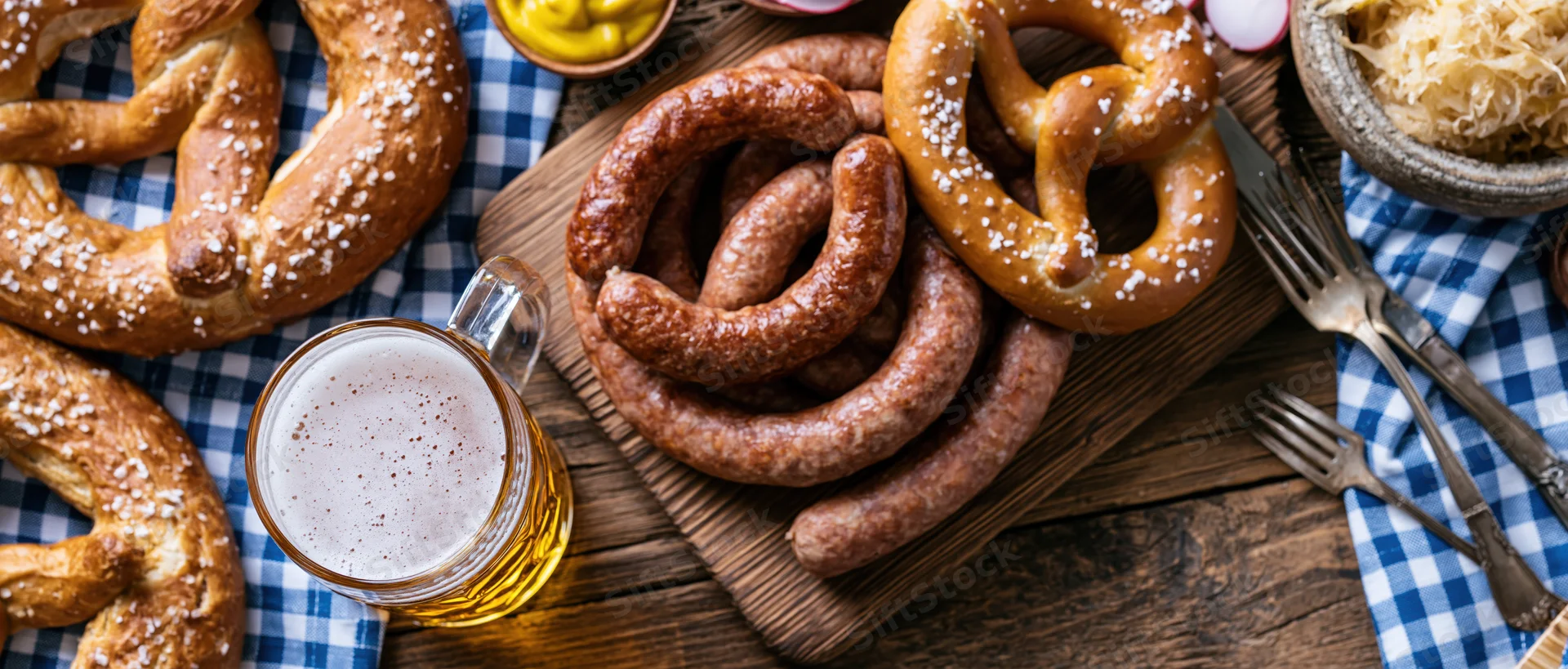 Overhead flat lay of bavarian oktoberfest food spread with sausages, pretzels, beer, and condiments on wooden table with checkered napkin