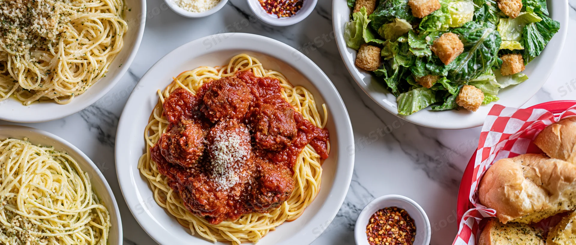Overhead flat lay of italian dinner spread with spaghetti meatballs salad bread on white marble table