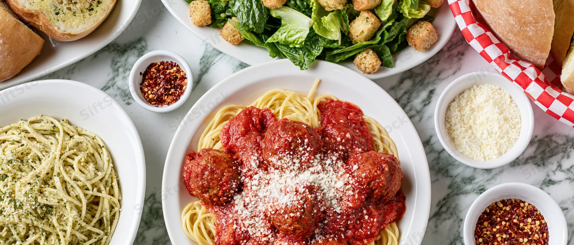 Spaghetti with meatballs and tomato sauce served on a white marble table alongside pesto pasta, salad, bread, and condiments