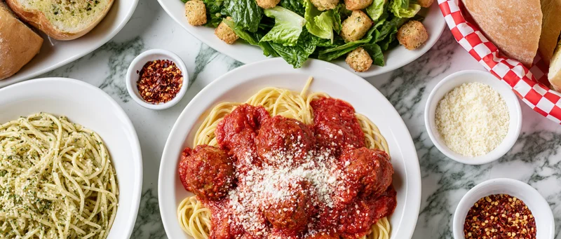 Spaghetti with meatballs and tomato sauce served on a white marble table alongside pesto pasta, salad, bread, and condiments