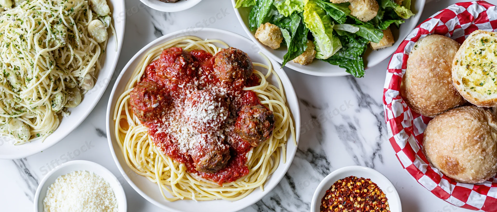 Spaghetti with meatballs and tomato sauce served on a white marble table alongside garlic bread, caesar salad, and parmesan cheese