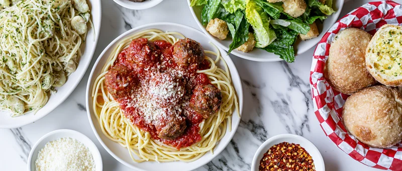 Spaghetti with meatballs and tomato sauce served on a white marble table alongside garlic bread, caesar salad, and parmesan cheese
