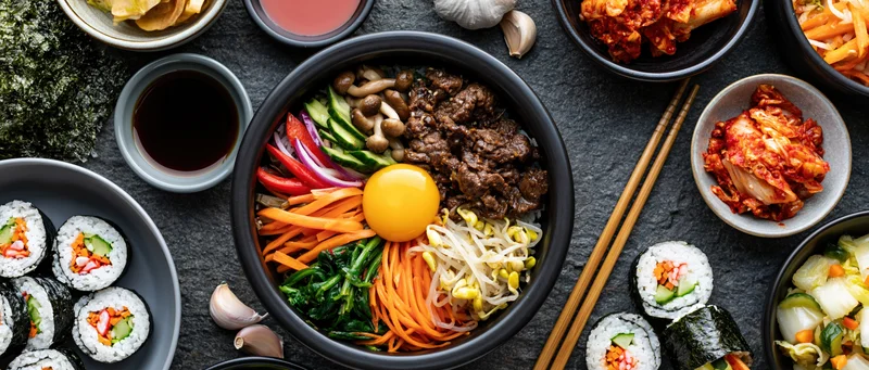 Overhead flat lay of bibimbap bowl with beef, egg, vegetables and kimchi surrounded by sushi and condiments on dark stone surface
