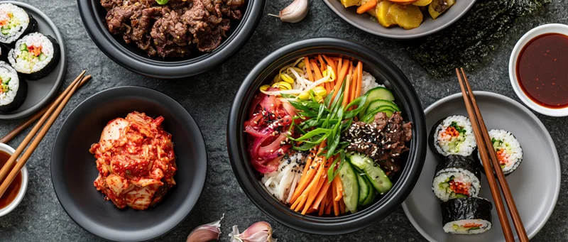 Overhead flat lay of bibimbap bowl with vegetables, beef, and kimchi surrounded by sushi, chopsticks, and soy sauce on dark surface