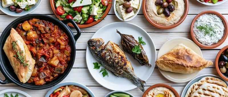 Greek mezze spread arranged overhead on a white wooden table with grilled fish, hummus, tzatziki, feta, olives, and