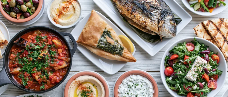 Overhead flat lay of a greek mezze spread on a white painted wooden table featuring grilled fish, hummus, tzatziki, spanakopita, salad, and pita bread