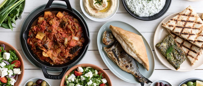 Overhead flat lay of a greek mezze spread on a white painted table with various dishes including grilled fish, pita, hummus, tzatziki, and salads