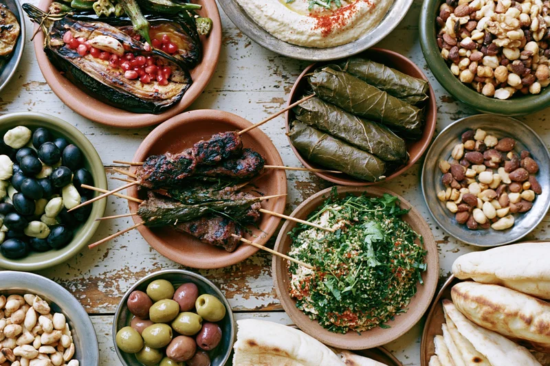 Overhead flat lay of a lebanese mezze spread on a worn wooden table with ceramic and metal bowls filled with hummus, tabbouleh, dolma, grilled eggplant, olives, nuts, pita bread, and skewered kebabs