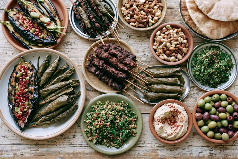 Overhead flat lay of a lebanese mezze spread on a worn wooden table with various dishes including kebabs, hummus, tabbouleh, and stuffed grape leaves