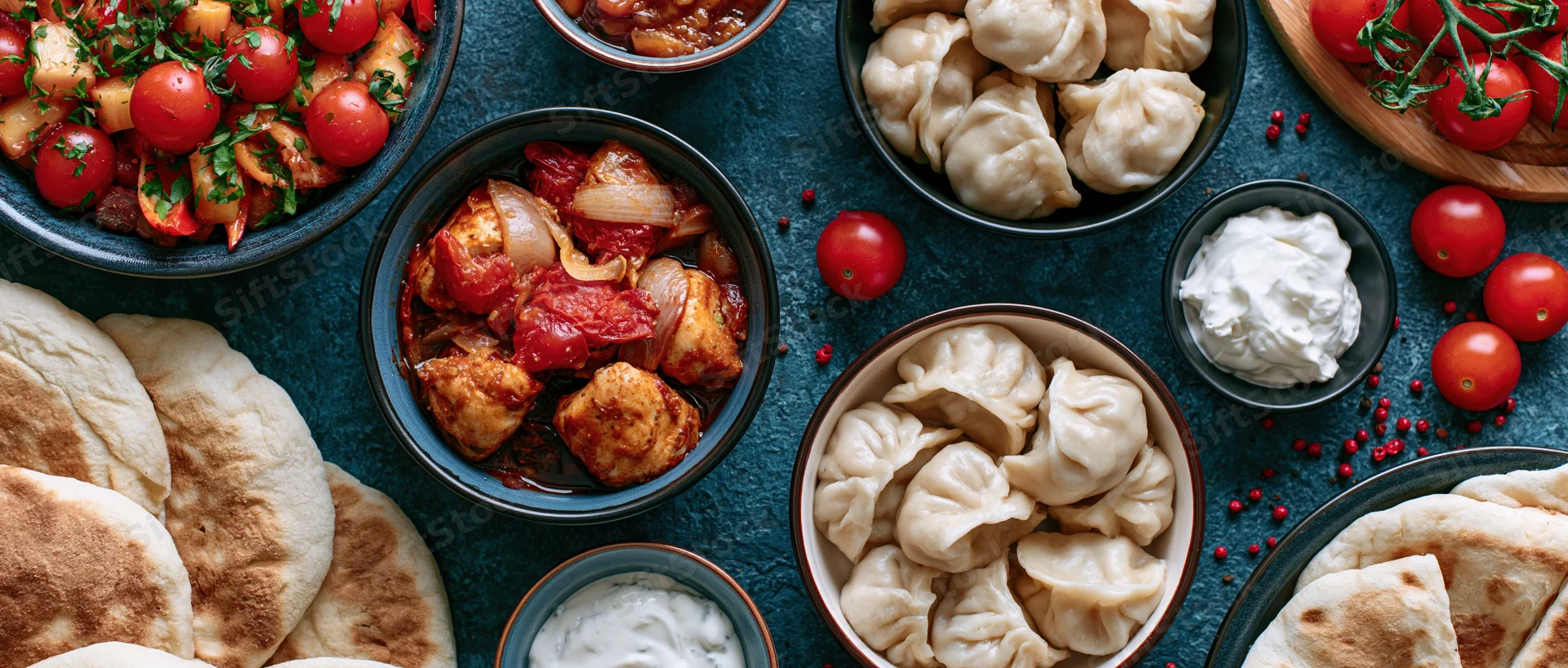 Overhead flat lay of mezze dishes including dumplings, flatbread, yogurt, and tomato-based sauces arranged on a dark textured surface