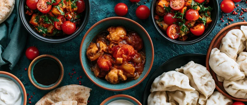 Overhead flat lay of mezze dishes with chicken stew, dumplings, flatbread, yogurt, and tomatoes on a dark blue textured surface