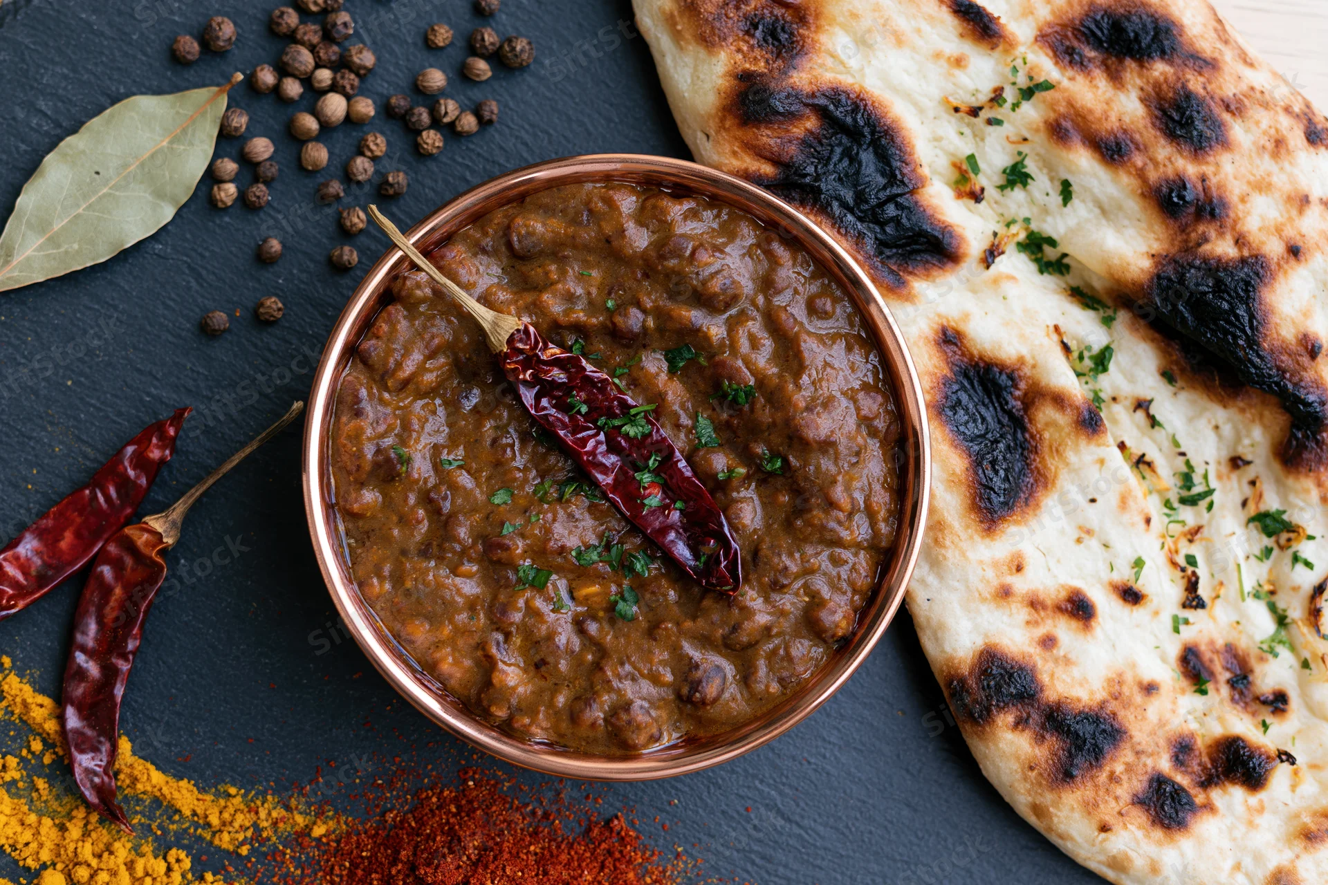 Copper bowl of dal makhani with naan and spices arranged on dark surface