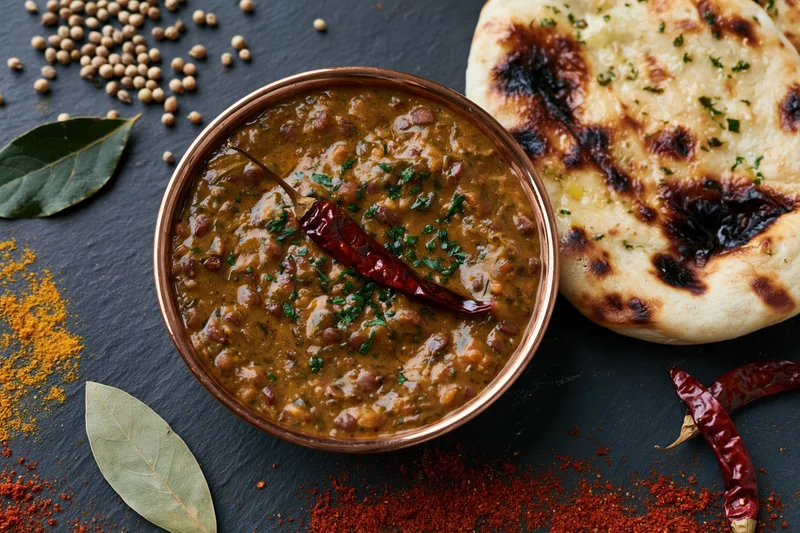 Copper bowl of dal makhani with naan and spices arranged on dark slate surface