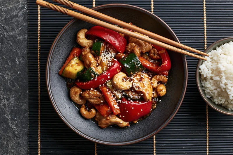 Stir fry with chicken and vegetables in a dark ceramic bowl with chopsticks and rice on a bamboo mat