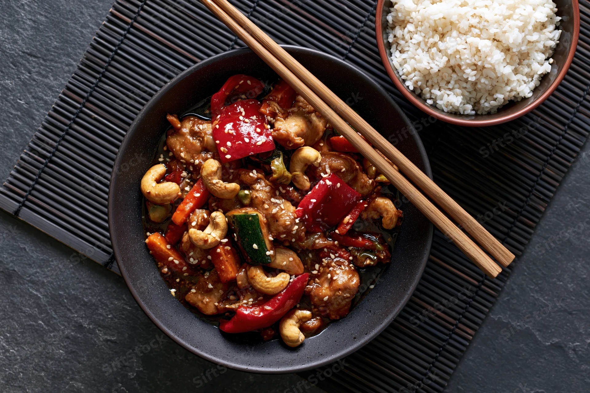 Stir fry with chicken and cashews in a dark ceramic bowl with chopsticks and rice on a bamboo mat