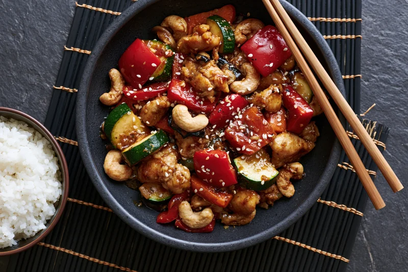 Stir fry with chicken, cashews, bell peppers, zucchini, and sesame seeds in a dark matte ceramic bowl with chopsticks and a side of white rice on a bamboo placemat
