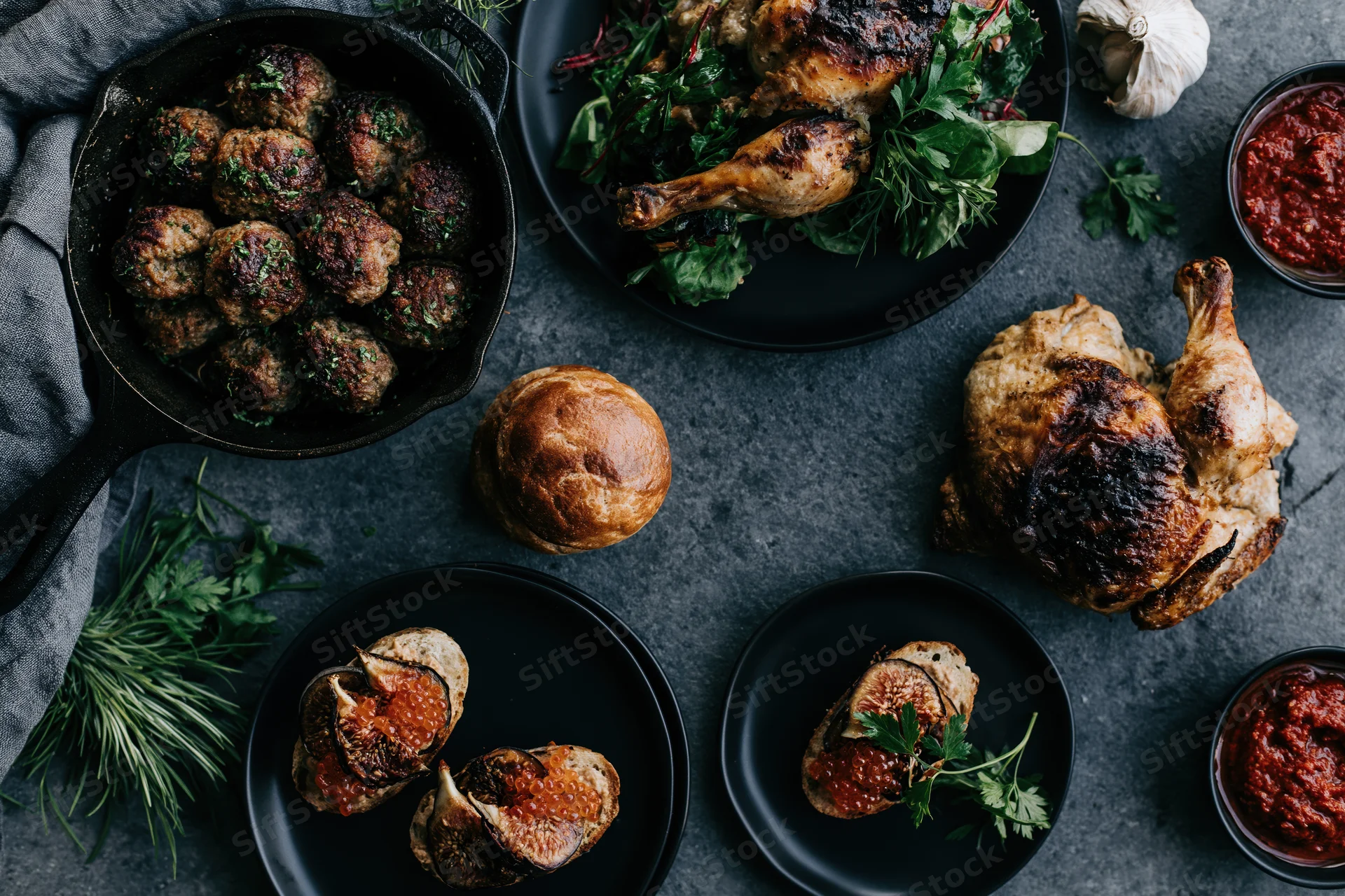 Overhead flat lay of a dinner spread with roasted chicken, meatballs, figs and herbs on a dark slate surface