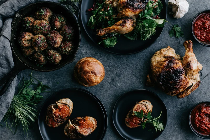 Overhead flat lay of a dinner spread with roasted chicken, meatballs, figs and herbs on a dark slate surface