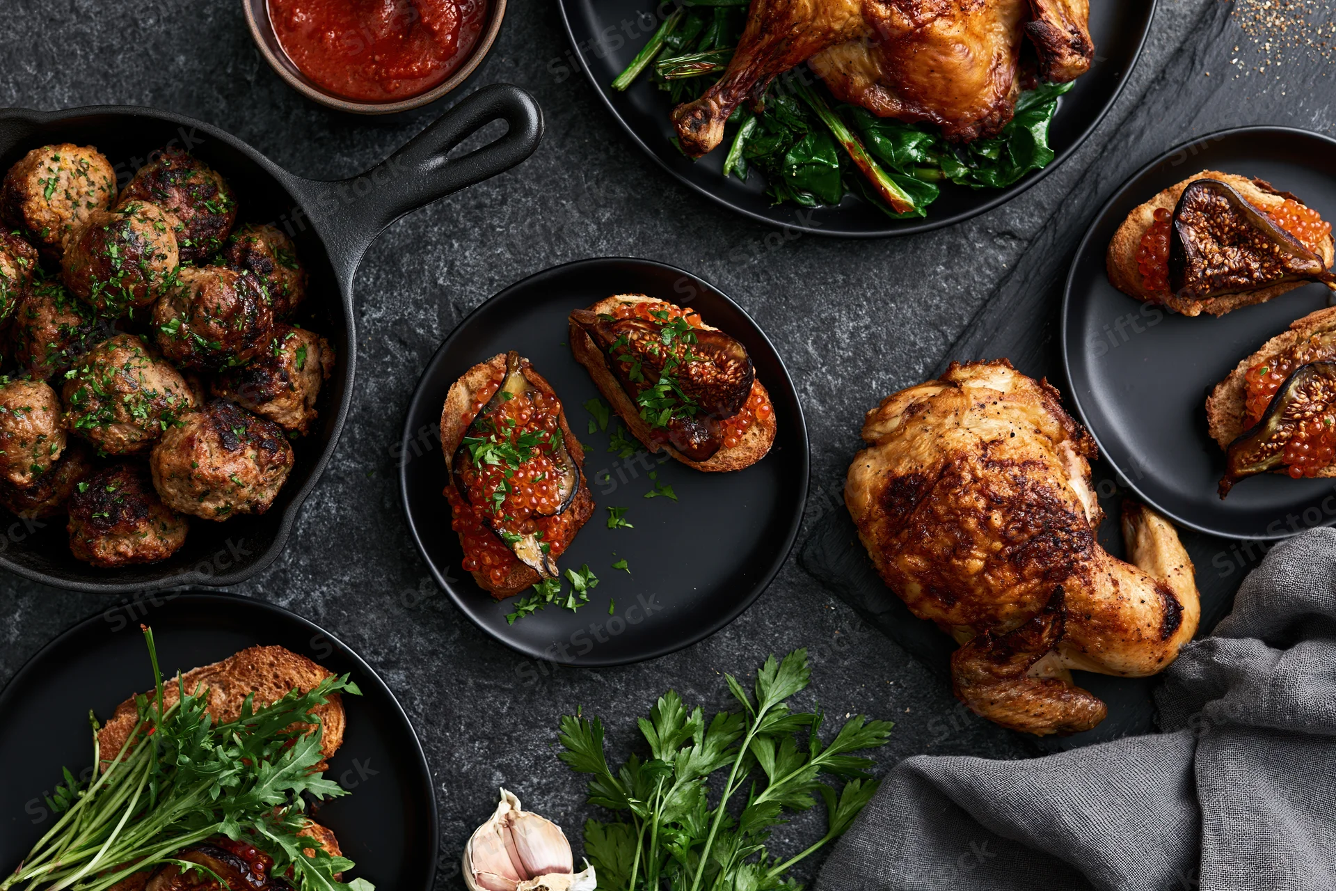 Overhead flat lay of a dinner spread with roasted chicken, meatballs, fig sandwiches and herbs on dark slate surface