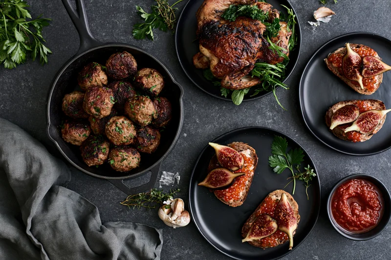Overhead flat lay of a dinner spread with meatballs, roasted chicken, fig toasts and herbs on a dark slate surface