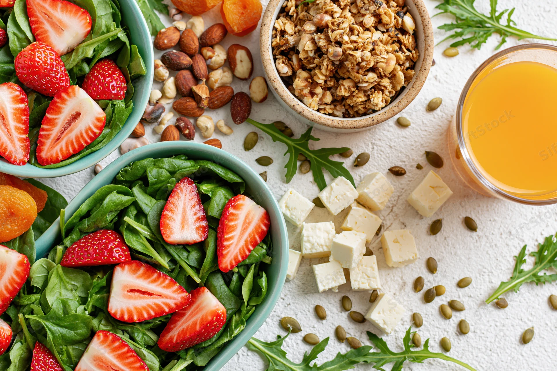 Overhead flat lay of a healthy breakfast spread with salads, granola, nuts, tofu, and orange juice on a white surface