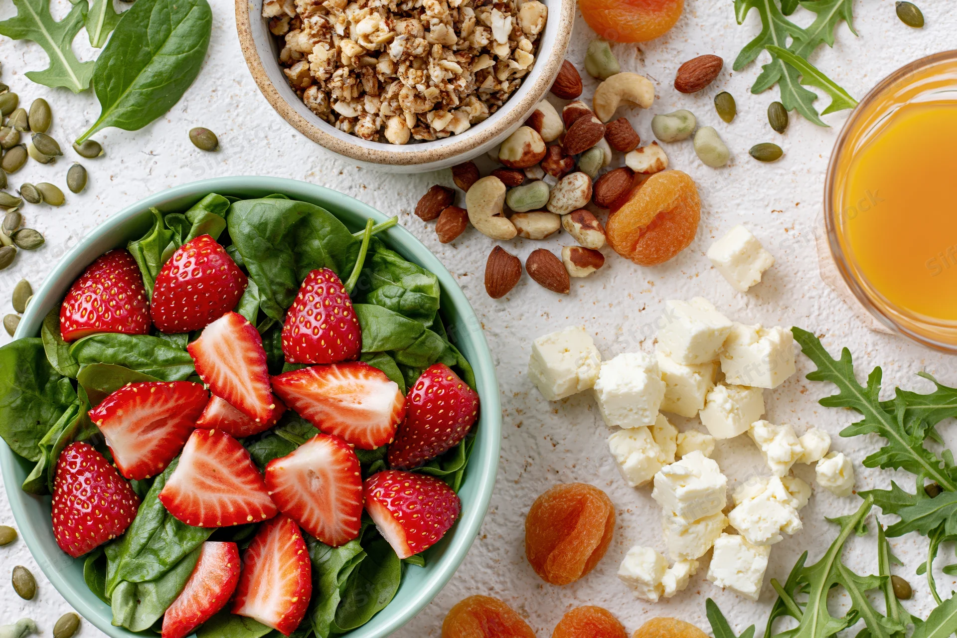 Overhead flat lay of a healthy breakfast spread with strawberries, spinach, granola, nuts, feta cheese, and orange juice on a white surface
