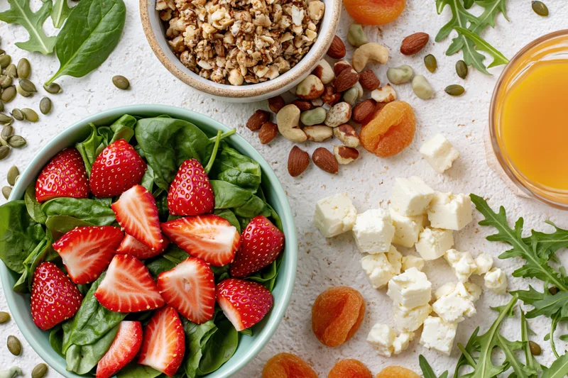 Overhead flat lay of a healthy breakfast spread with strawberries, spinach, granola, nuts, feta cheese, and orange juice on a white surface
