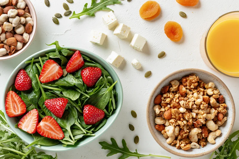 Overhead flat lay of a healthy breakfast spread with salad, granola, nuts and orange juice on a white surface