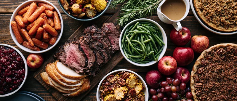 Overhead flat lay of a holiday feast spread across a dark wooden table with roast beef, turkey, vegetables, fruit, pies and gravy
