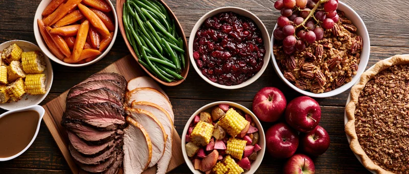 Overhead view of a thanksgiving feast spread on a dark wooden table with turkey, beef, corn, cranberry sauce, pie, apples, grapes, and side dishes